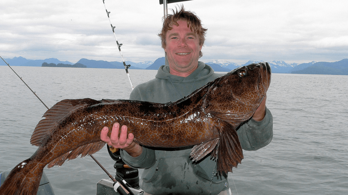 A smiling man holds a large fish while standing on a boat in a scenic water landscape.