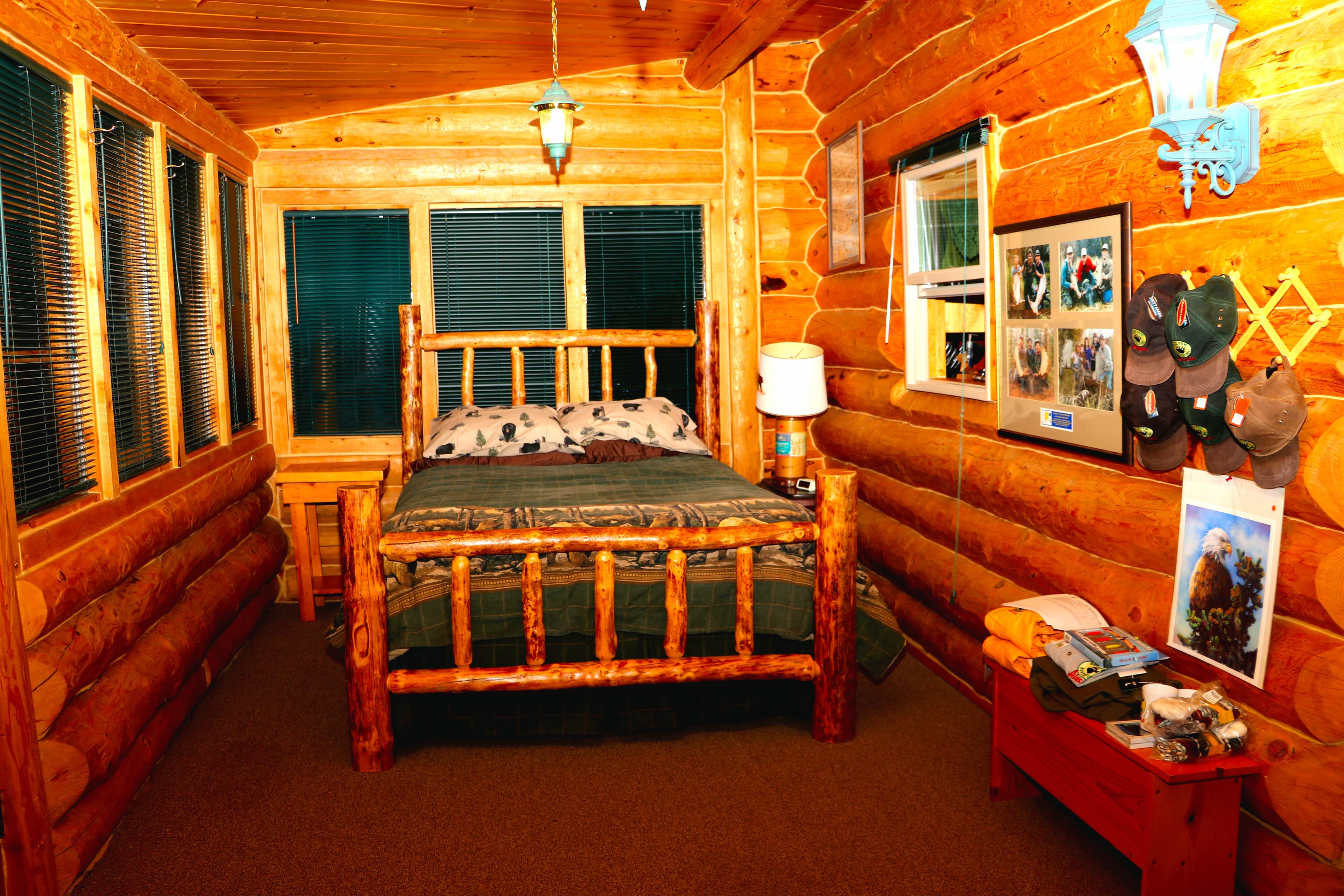 The room features a wooden-framed queen bed with a dark green comforter and patterned pillows in muted tones. A small wooden nightstand with a lamp featuring a white shade sits beside the bed.