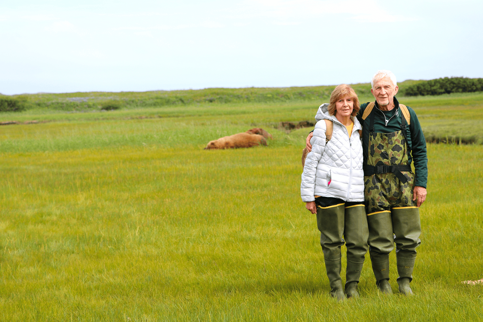 A couple stands together in a grassy field, with a bear resting in the background.