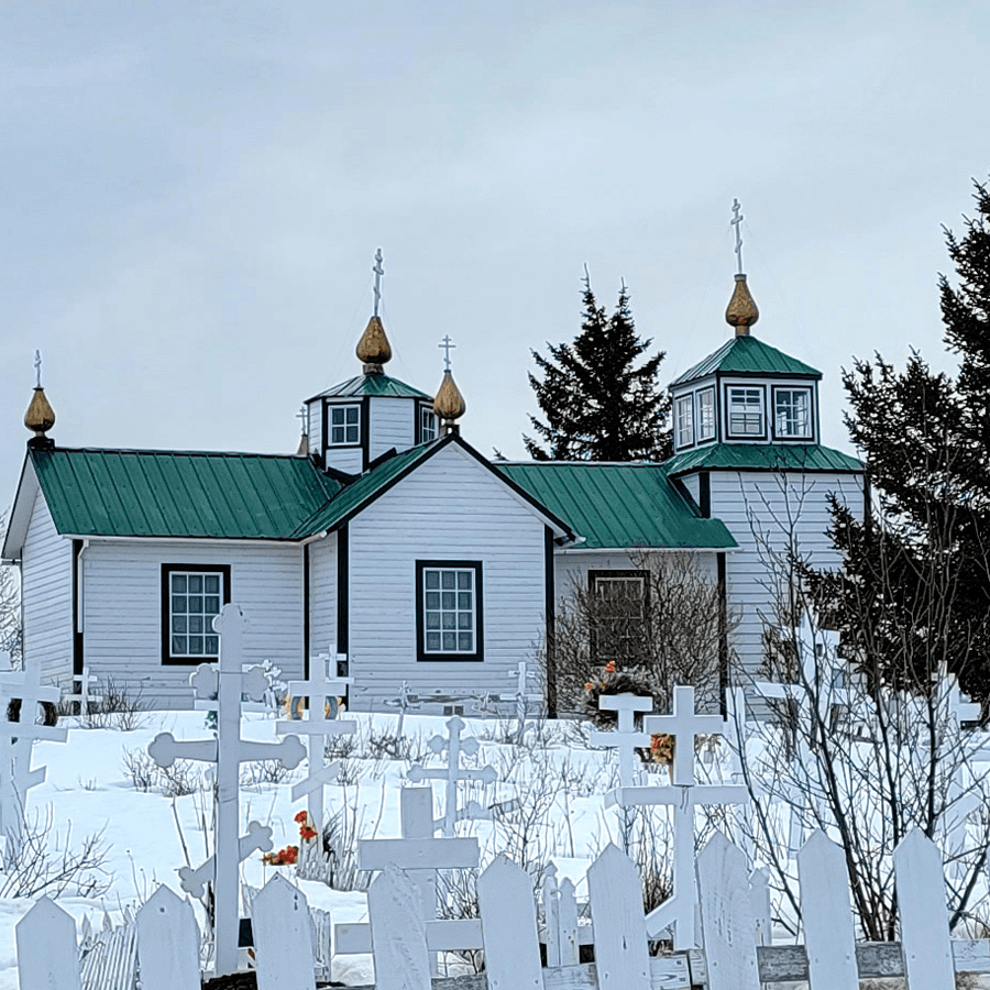 External view of a cloudy day showing a white Russian church with a green roof and white crosses marking graves in the foreground, with snow on the ground.