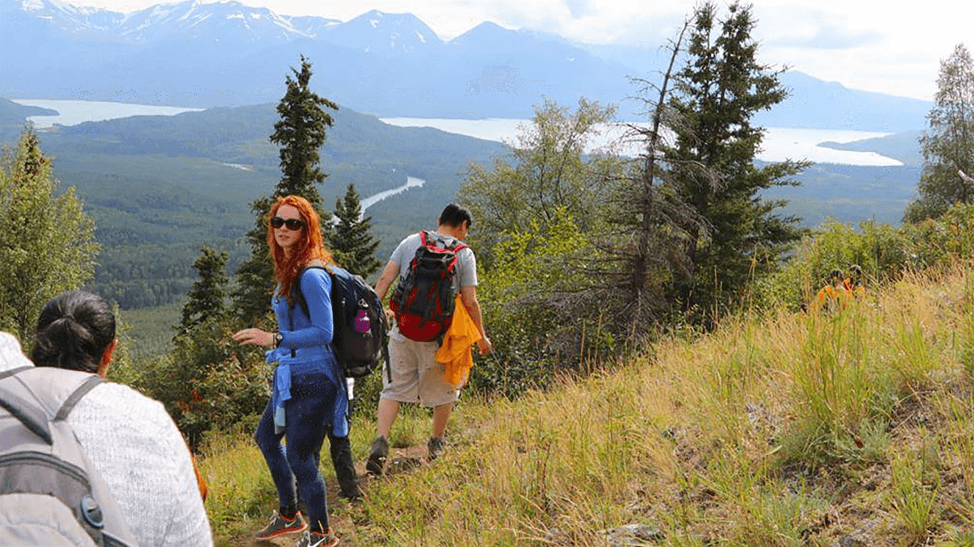 Group of people including a redheaded woman with sunglasses hiking in the grass on the side of a mountain, with Alaska mountains in the background