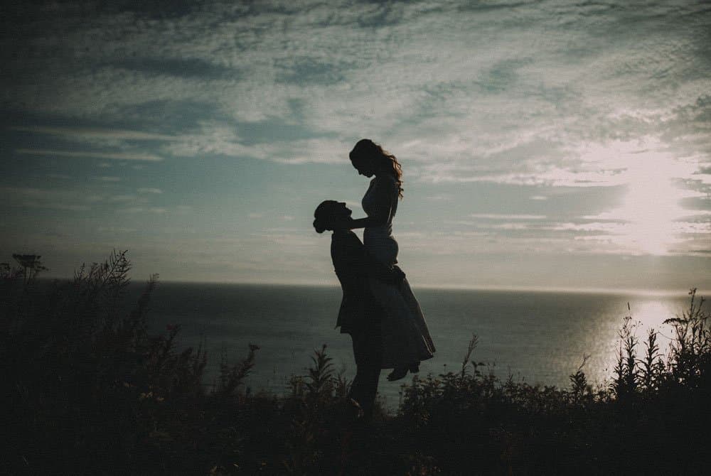 A silhouetted couple shares a romantic moment against a coastal sunset.