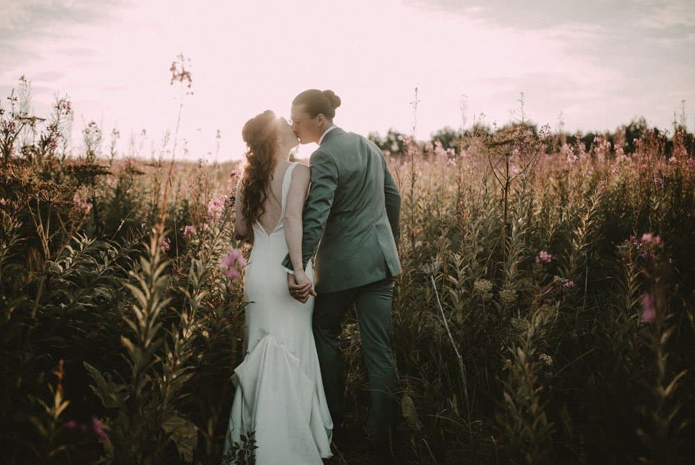 A couple in wedding attire shares a kiss in a field of flowers at sunset.