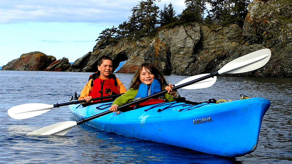 A child and an adult are kayaking on a calm body of water with rocky cliffs in the background.