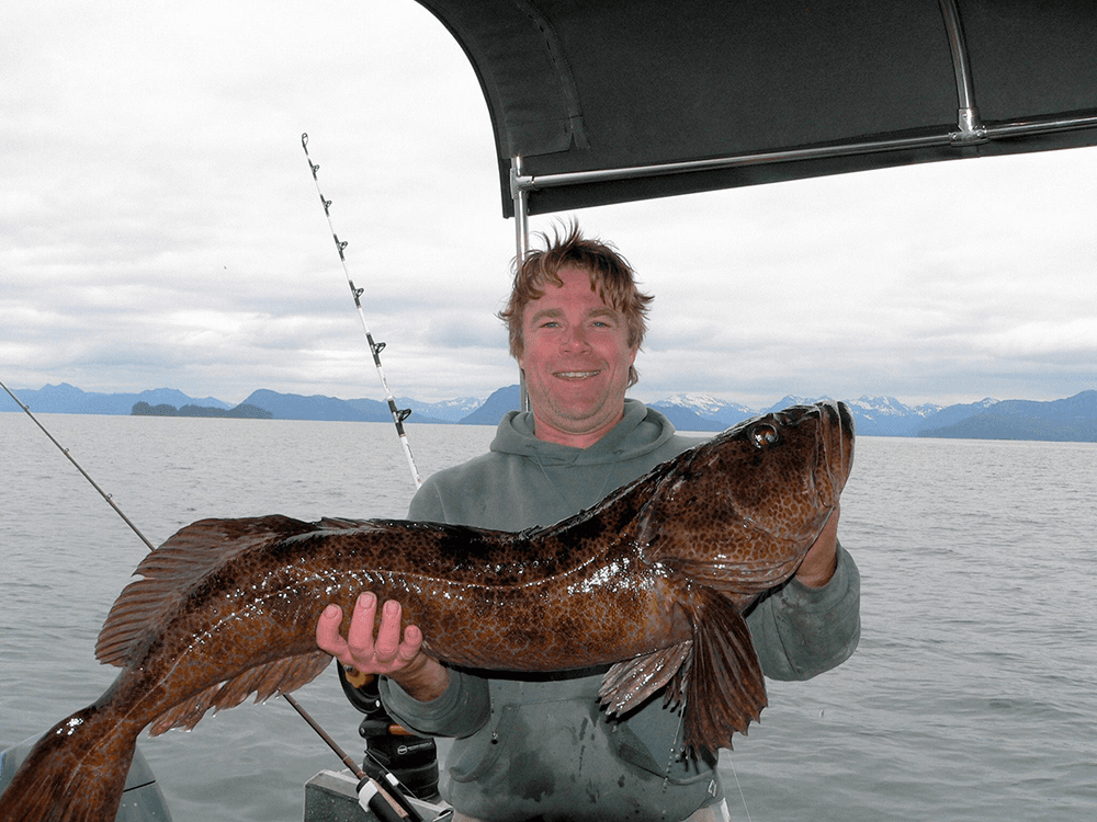 A person holds a large fish while standing on a boat amid a scenic backdrop of mountains and water.