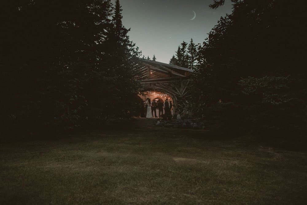 A group of people stand at the entrance of a rustic, illuminated building surrounded by trees at dusk.