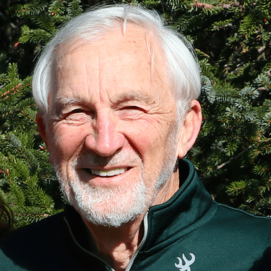 Smiling older man with gray hair and a beard, wearing a dark jacket, in front of green foliage.