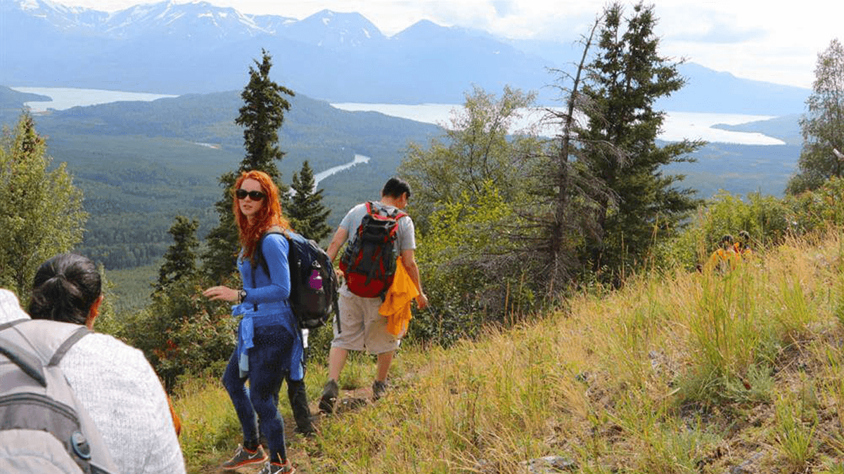 A group of hikers navigates a lush mountainside with a scenic view of a river and mountains in the background.