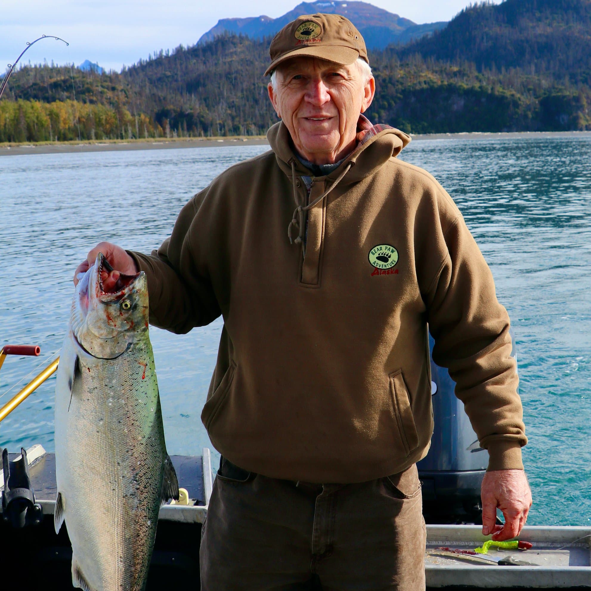 An older man smiles while holding a large fish against a scenic waterfront backdrop.