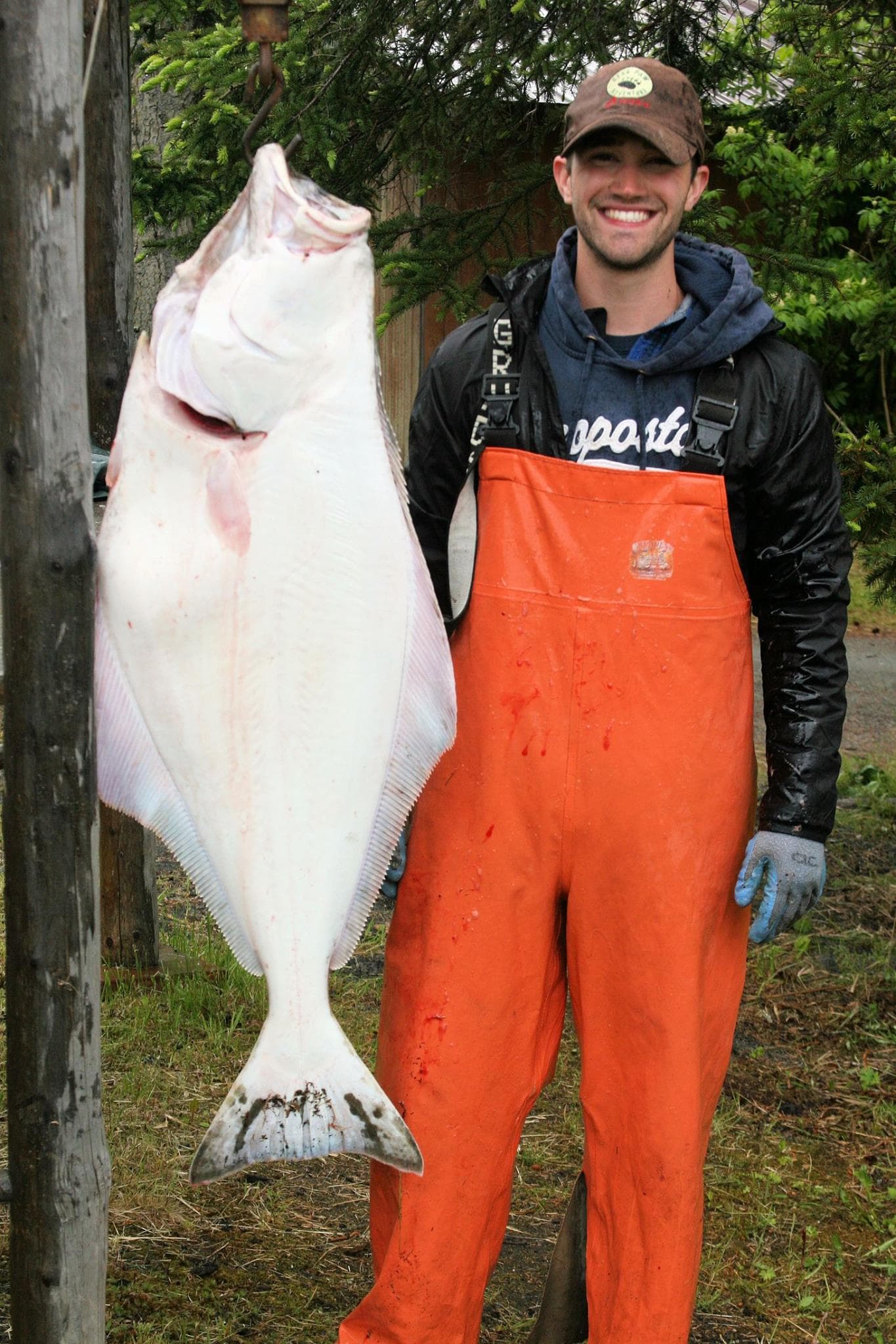A smiling person in orange overalls holds a large halibut fish on a hook.