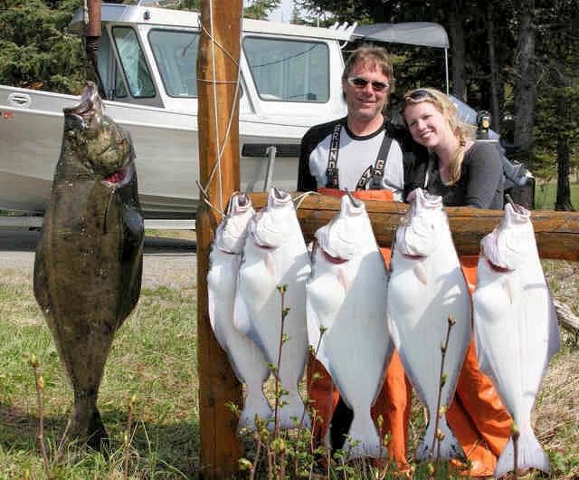 A man and woman pose proudly beside a display of freshly caught fish, including a large halibut, with a boat in the background.