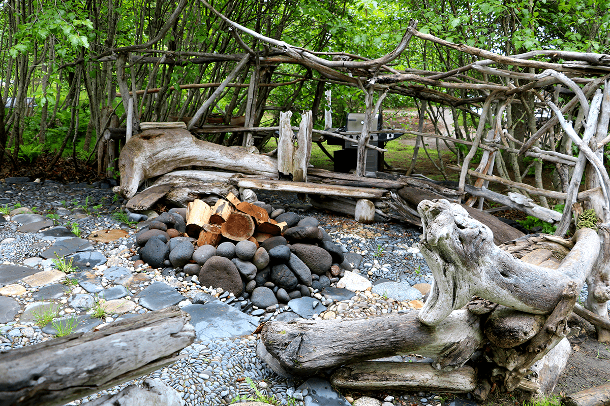 A rustic outdoor seating area surrounded by trees, featuring a stone fire pit with logs.