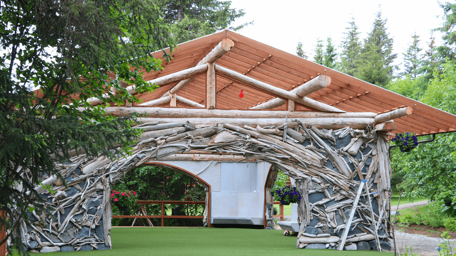 A rustic wooden structure with a log archway surrounded by greenery and colorful flower pots.