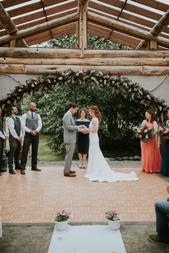 A bride and groom exchange vows under a floral arch during an outdoor wedding ceremony.