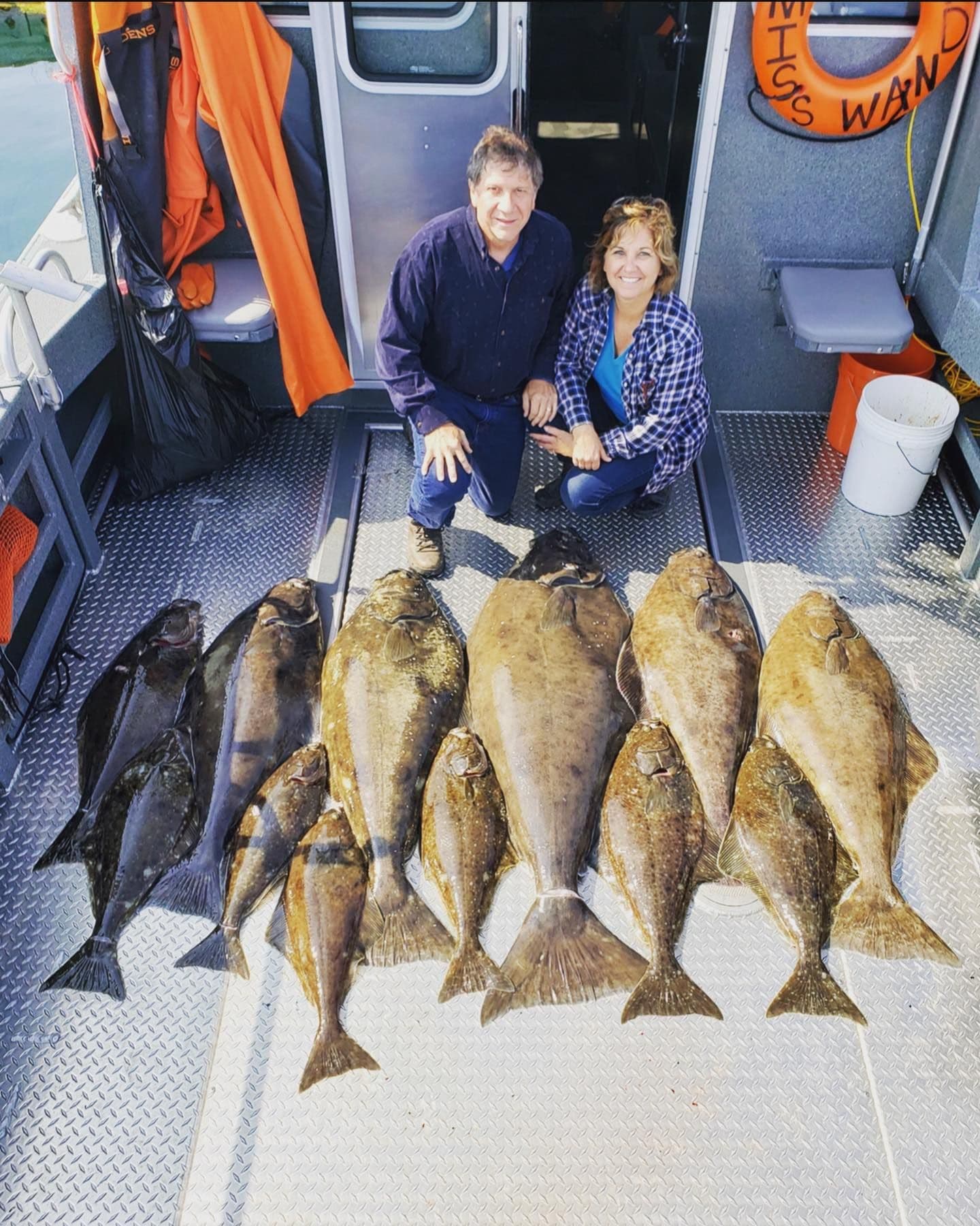A man and a woman pose on a boat with a large catch of flatfish displayed on the deck.
