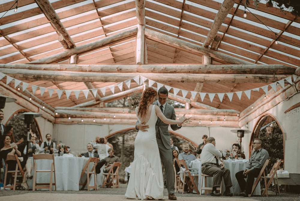 A bride and groom share a romantic dance under a rustic wooden structure while guests watch and celebrate.