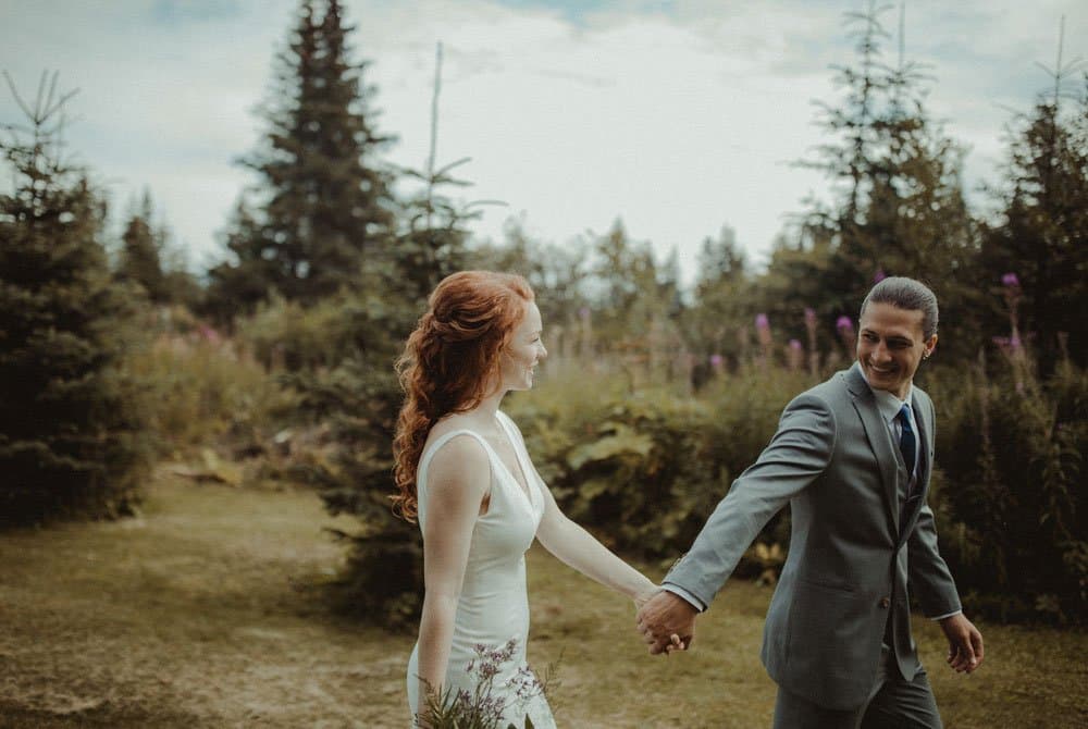 A bride and groom walk hand in hand through a lush, green landscape.