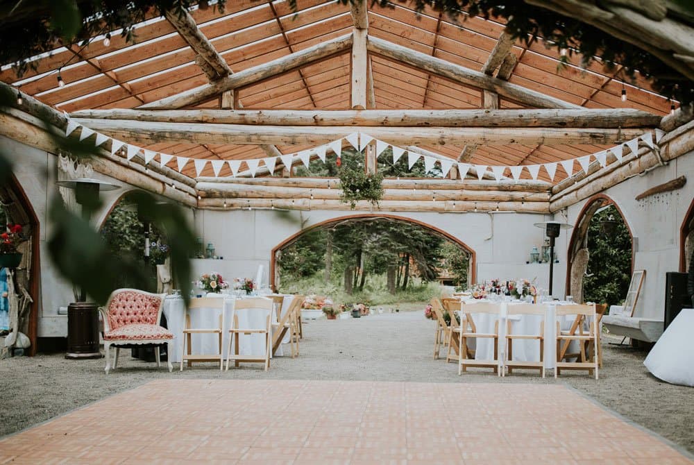 A beautifully decorated event space featuring wooden beams, round tables, and bunting, set in a natural setting.