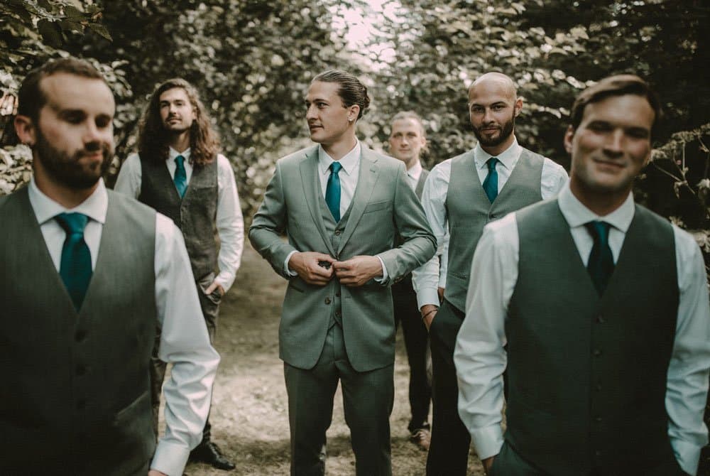 A group of six men in formal outfits stands together in a wooded setting.