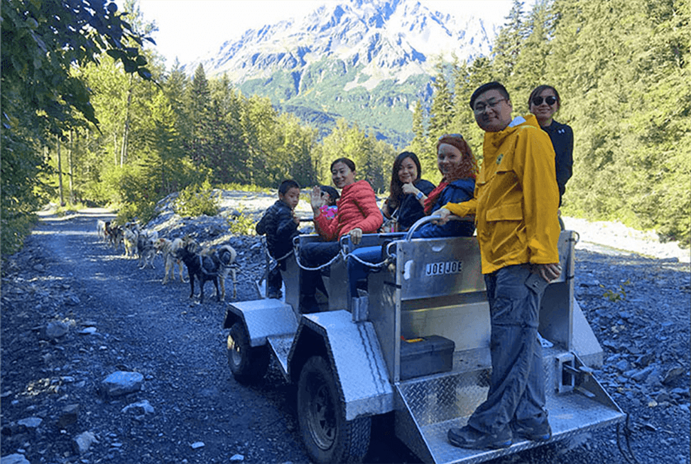 A group of people sits on a vehicle beside a dog team on a gravel path, surrounded by mountains and trees.