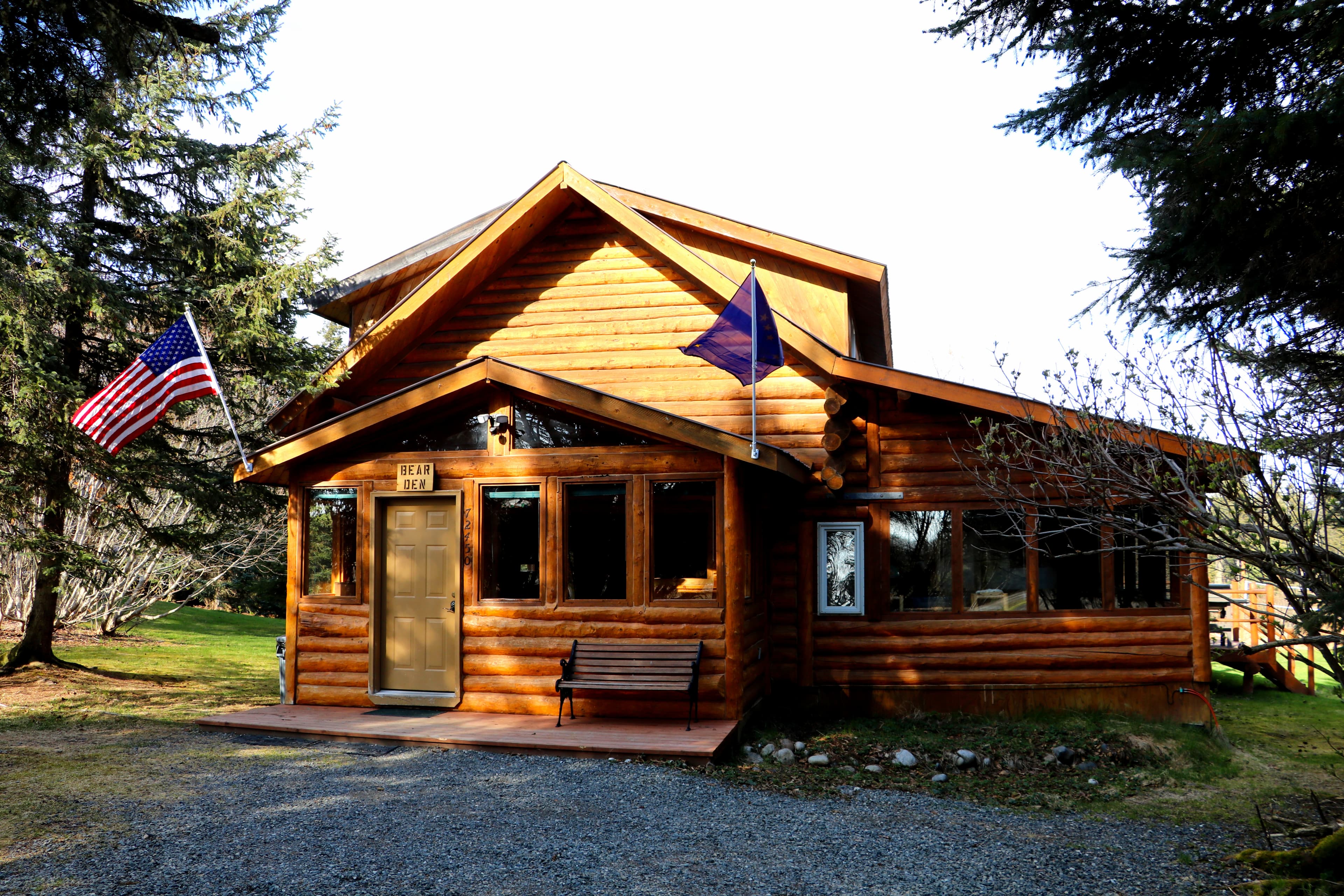 Street view of a charming two-story log cabin surrounded by evergreens under a bright sky.