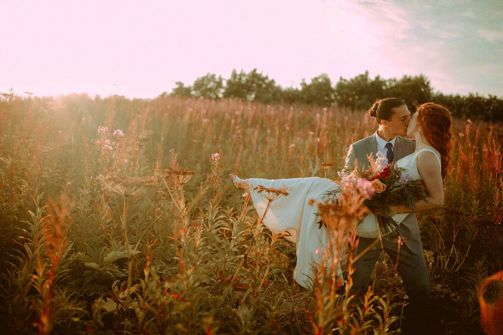 A couple kisses in a sunlit field, surrounded by tall grasses and wildflowers.