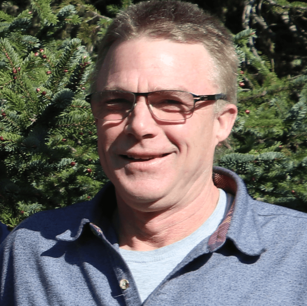 A smiling man with glasses stands outdoors in front of greenery.