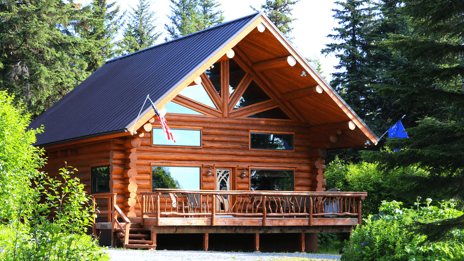 A rustic log cabin with a black metal roof, surrounded by lush greenery and flag poles.