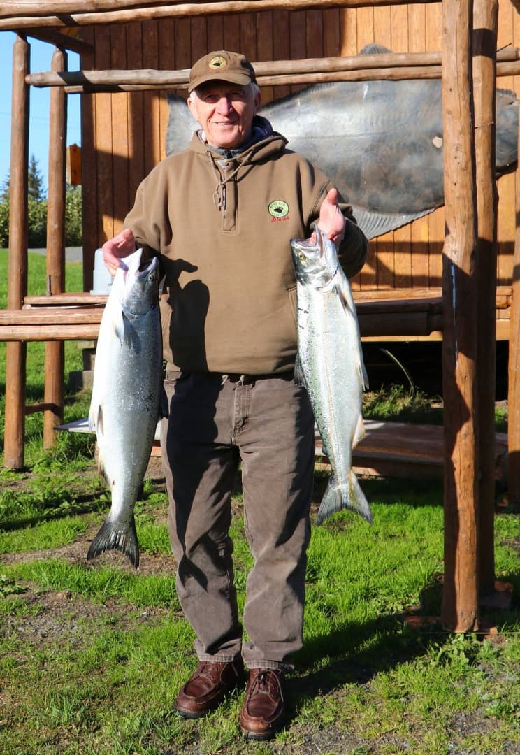 An older man holds two large fish outside a wooden structure.