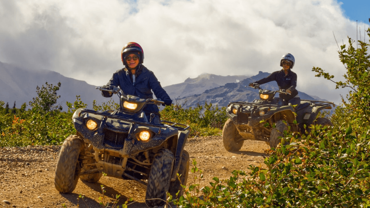 Two girls wearing helmets and sunglasses, riding ATVs on a dirt road with clouds and mountains in the background