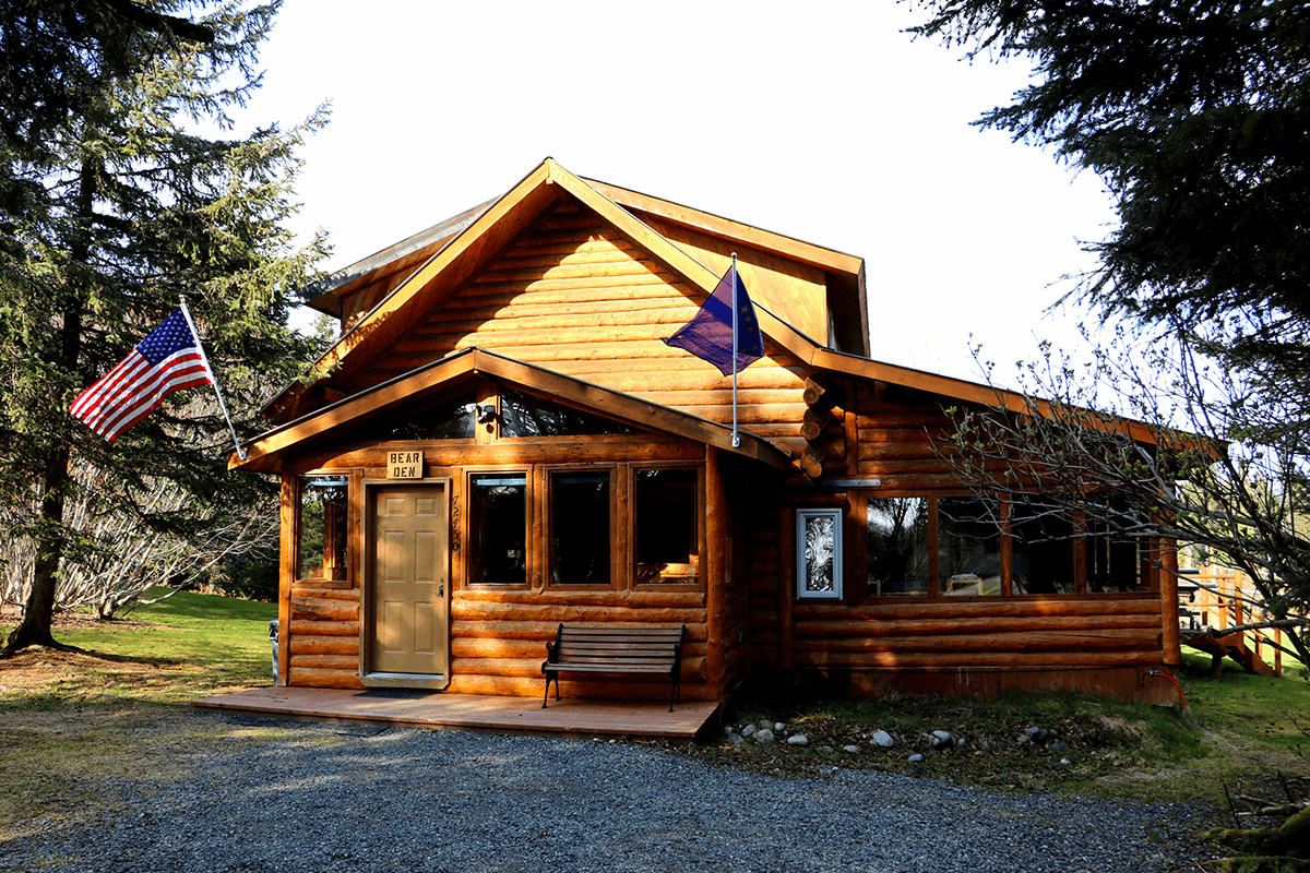 A log cabin set between two evergreen trees with a USA flag and an Alaska state flag affixed to the outside of the cabin.