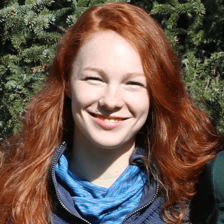 A smiling person with red hair stands in front of greenery.