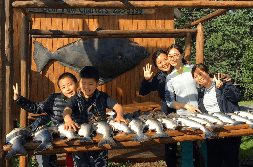 A group of four adults and children joyfully pose with freshly caught fish on a wooden table.