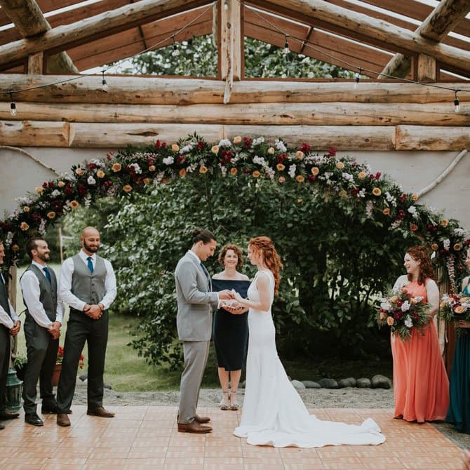 Bride and groom stand in front of a woman officiant under large floral arch holding hands as they prepare to say their wedding vows