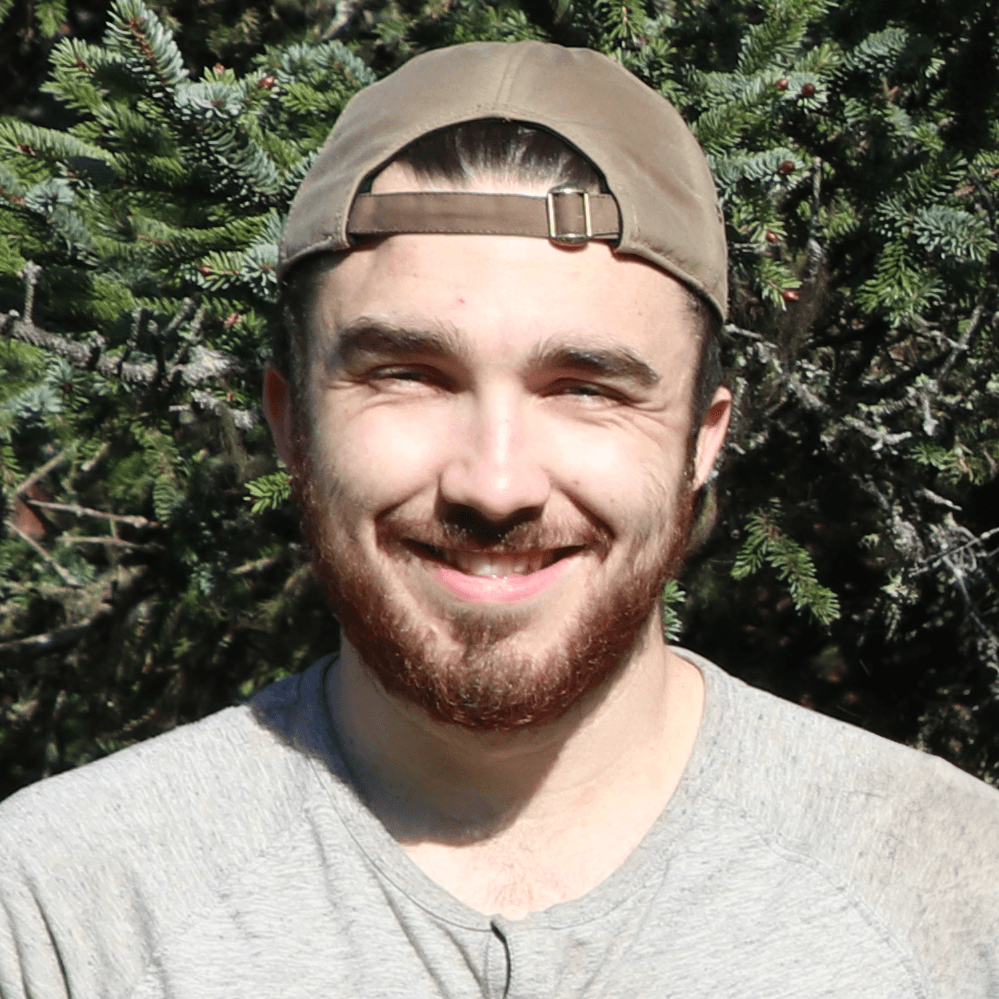 A smiling young man with a beard and cap stands in front of greenery.