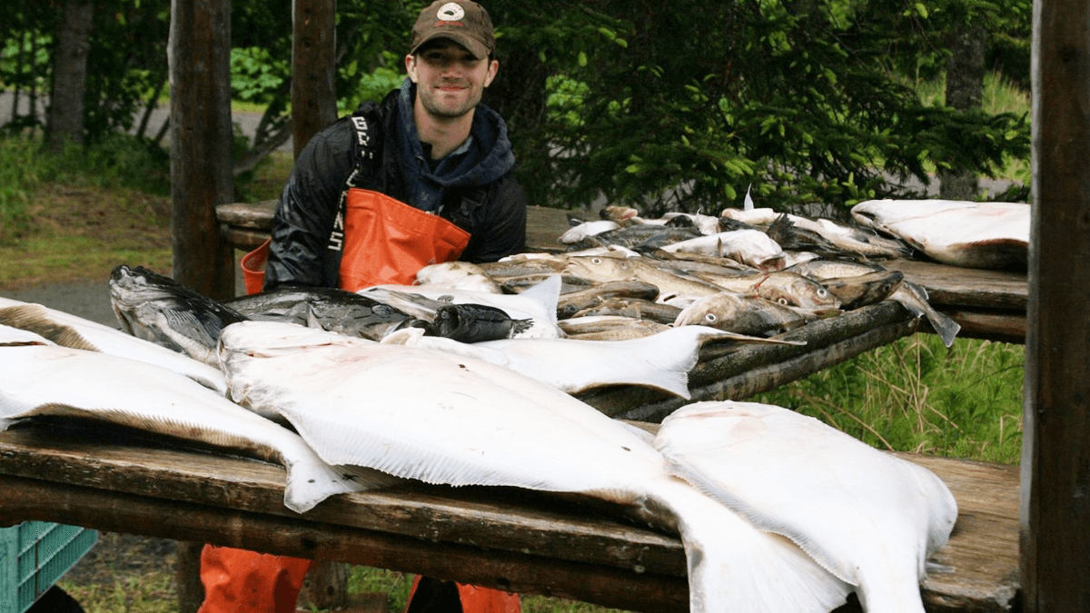 A smiling fisherman stands in front of a table covered with various freshly caught fish.