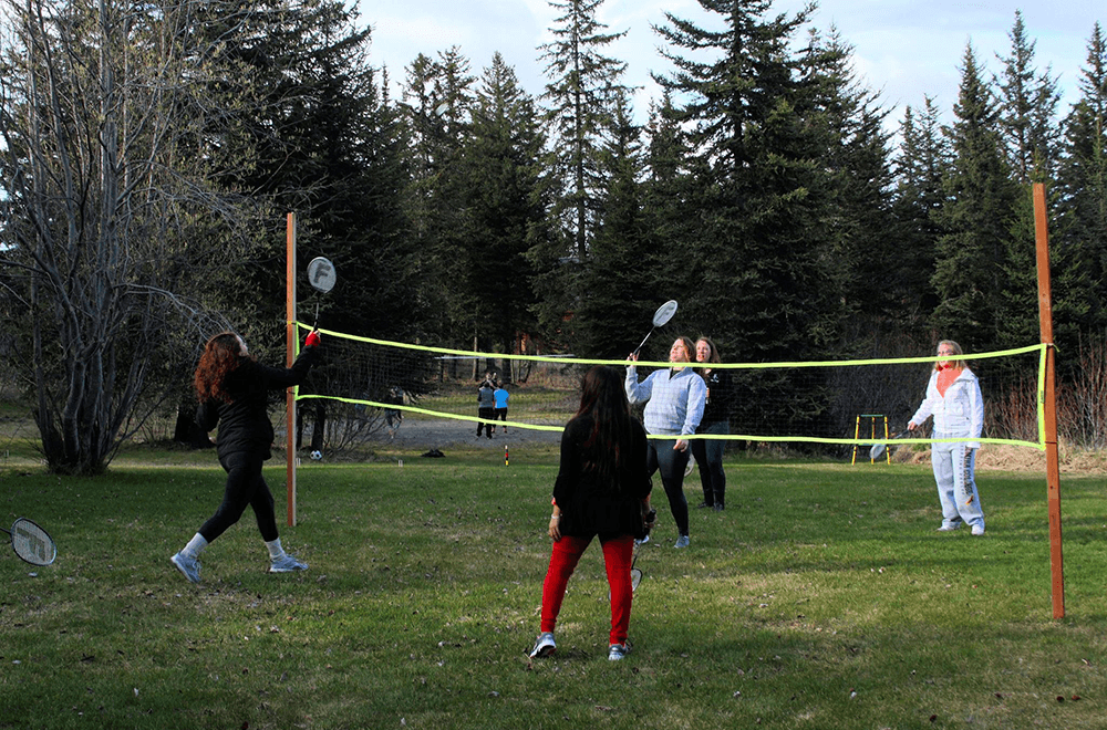 A group of people plays badminton outdoors under a clear sky, surrounded by trees.