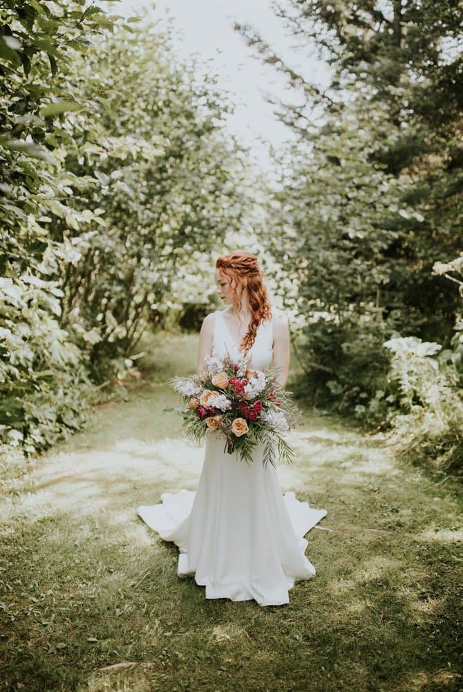 A bride in a white gown stands on a lush path, holding a colorful bouquet surrounded by greenery.