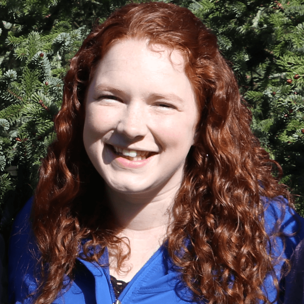 A smiling person with long, curly red hair, wearing a blue jacket, stands in front of evergreen trees.