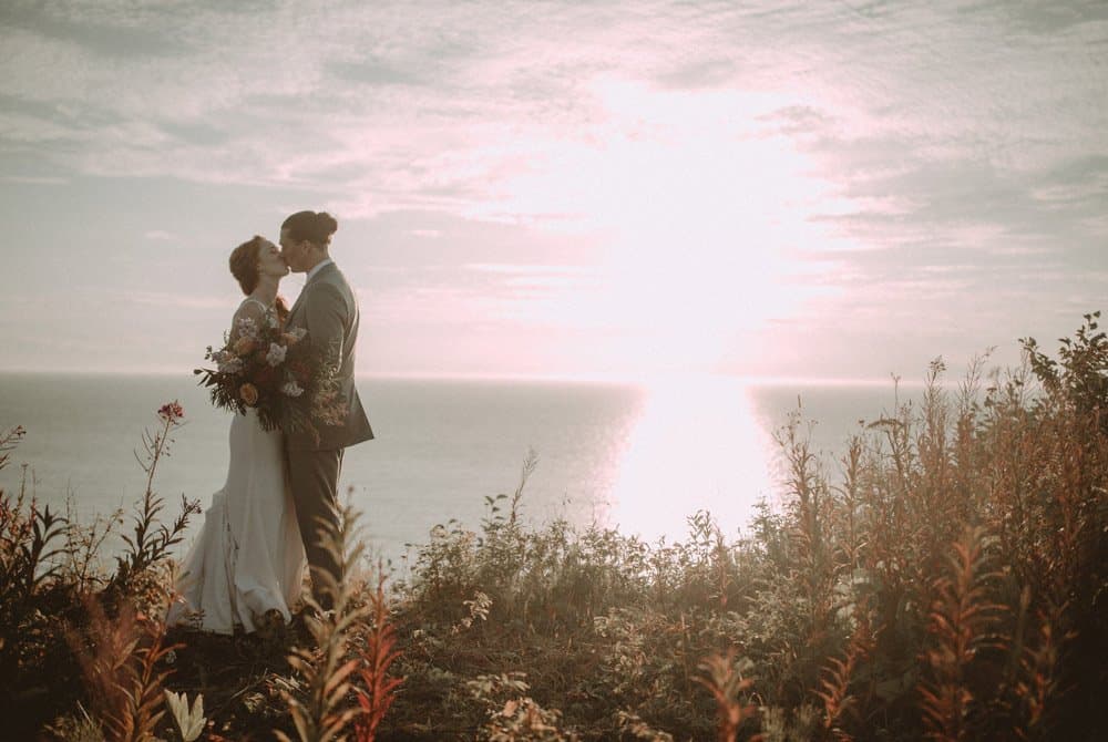 A couple shares a kiss against a sunset over the ocean, surrounded by wildflowers.