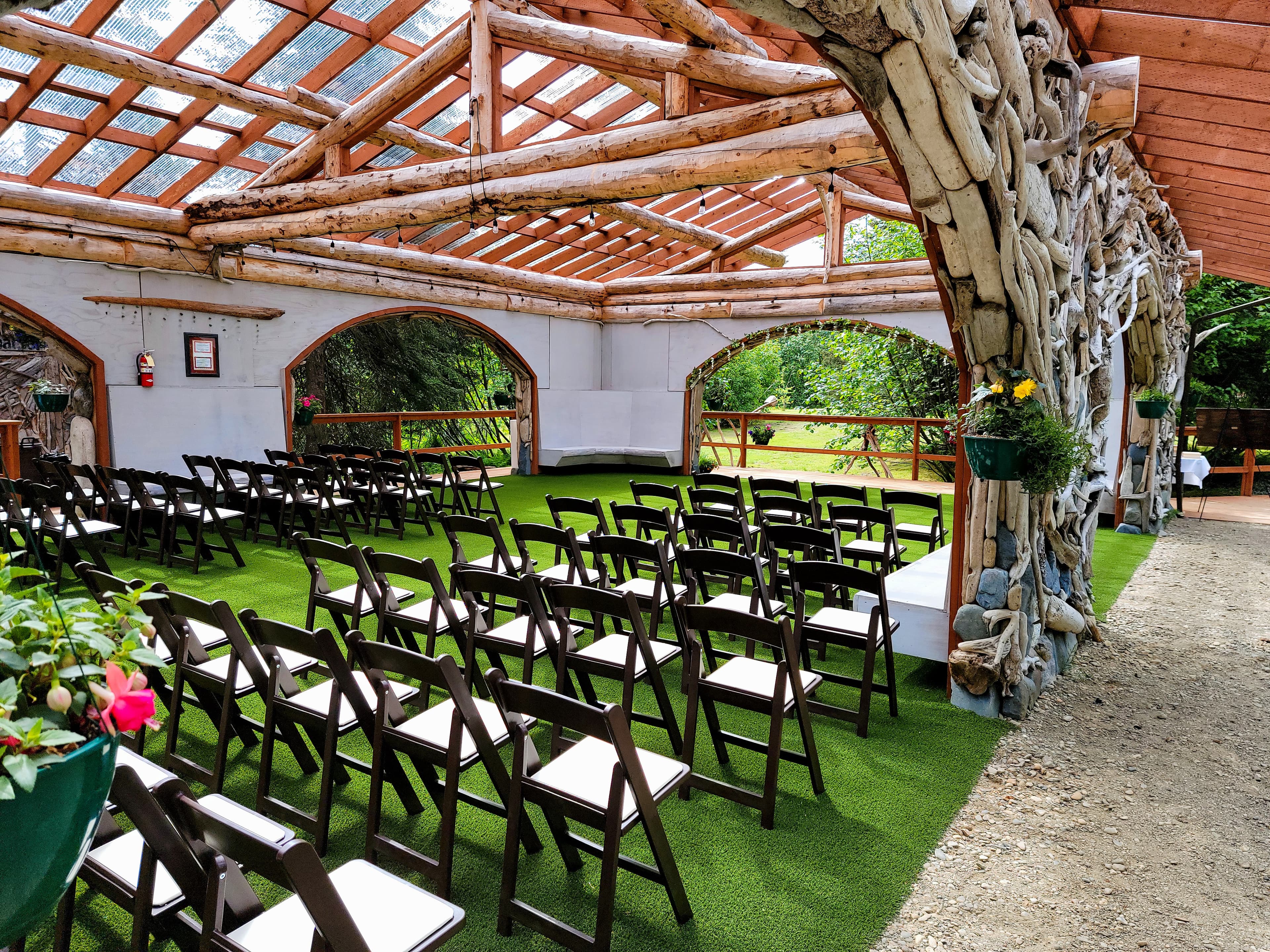 An open, rustic event space with wooden benches and a grassy floor, surrounded by natural light and greenery.