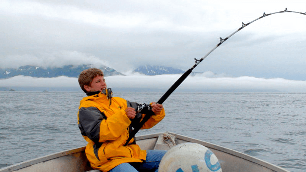 A young boy in a yellow rain jacket struggles to reel in a fishing rod while sitting in a boat on a cloudy day.