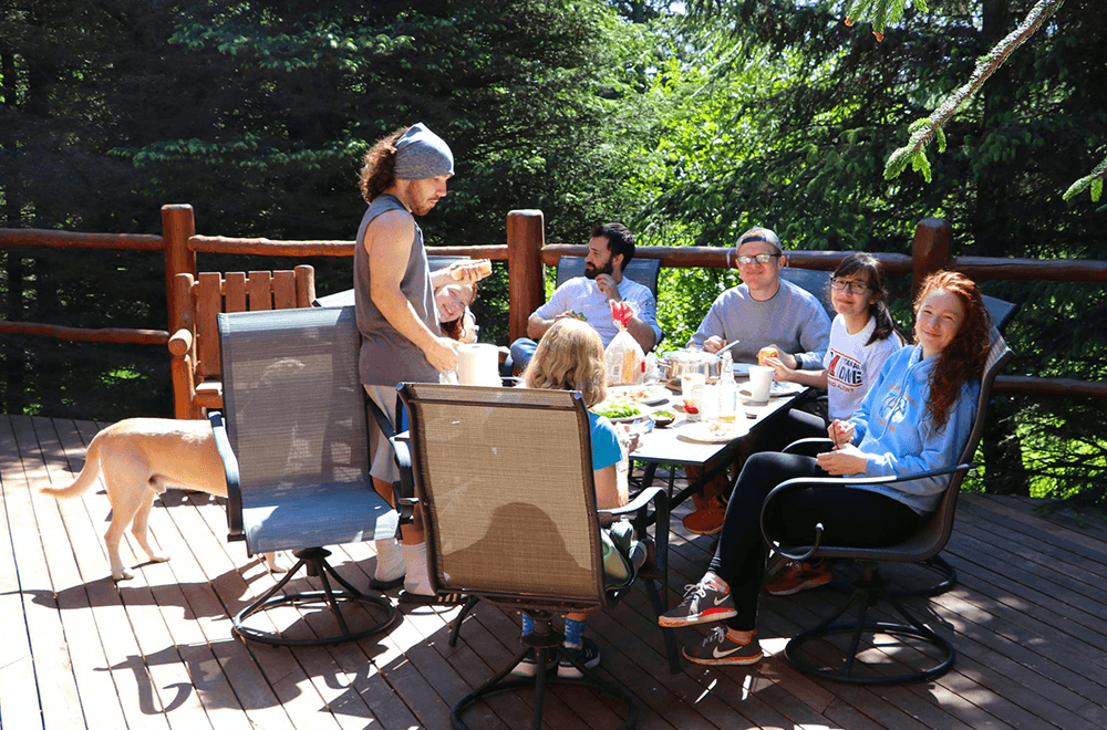 A group of people enjoy a meal together on a sunny deck surrounded by trees.