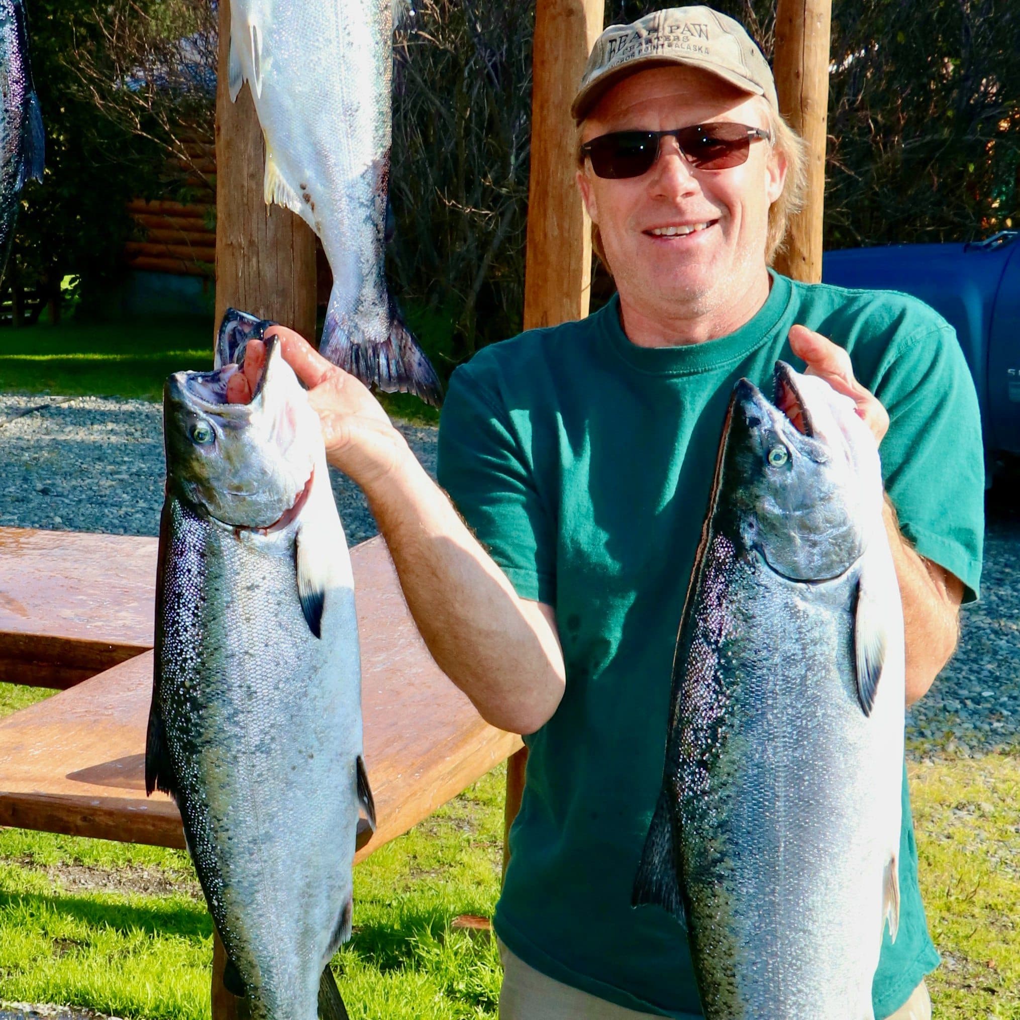 Outdoor image of a man with baseball cap and sunglasses holding up two large fishes, one in each hand, that he caught