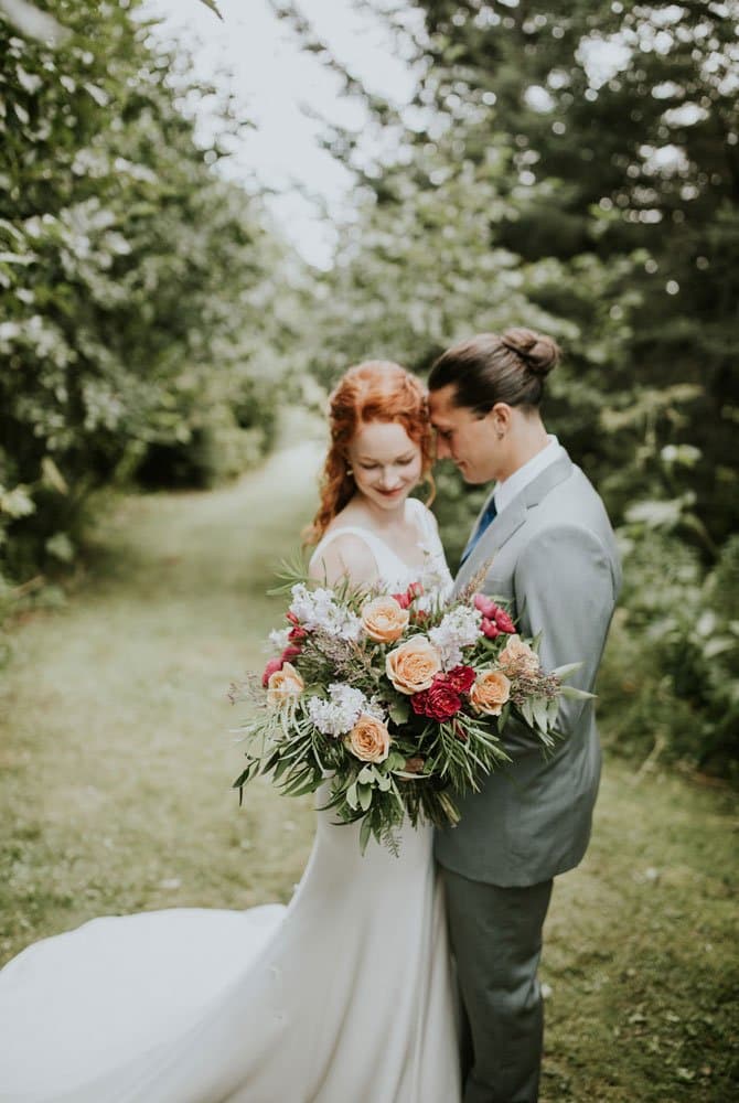 A bride and groom embrace in a lush, green pathway while the bride holds a colorful floral bouquet.