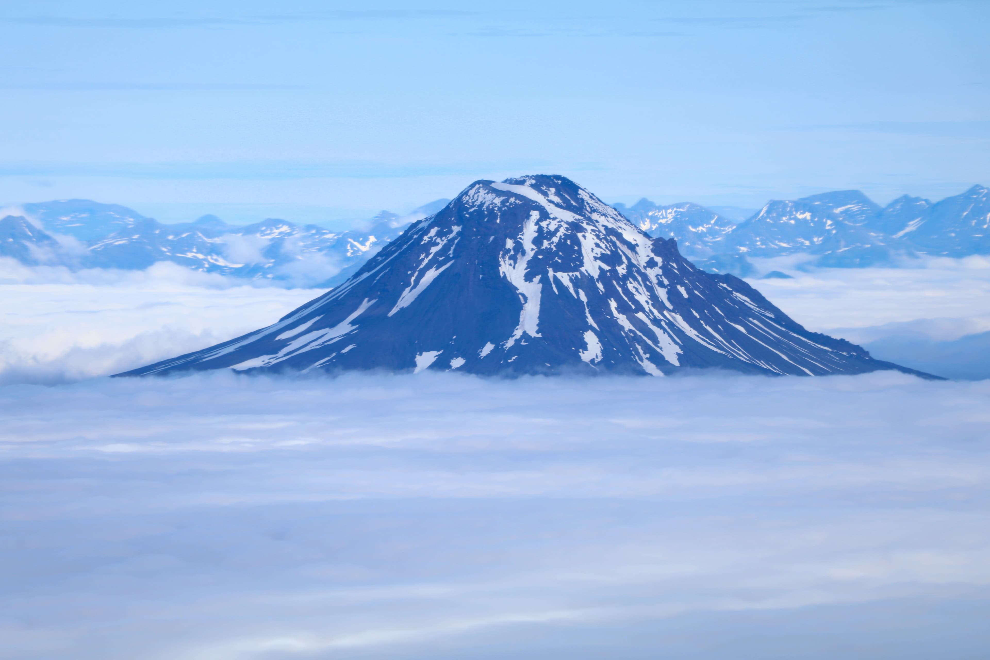 A snow-capped volcano rises above a blanket of clouds against a clear blue sky.