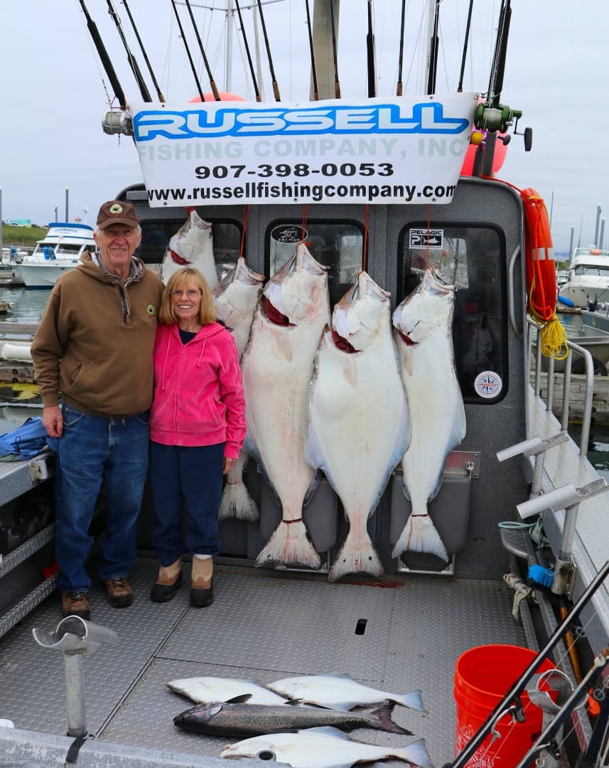 A man and a woman stand on a boat beside several large fish and fishing gear, with a sign for Russell Fishing Company in the background.