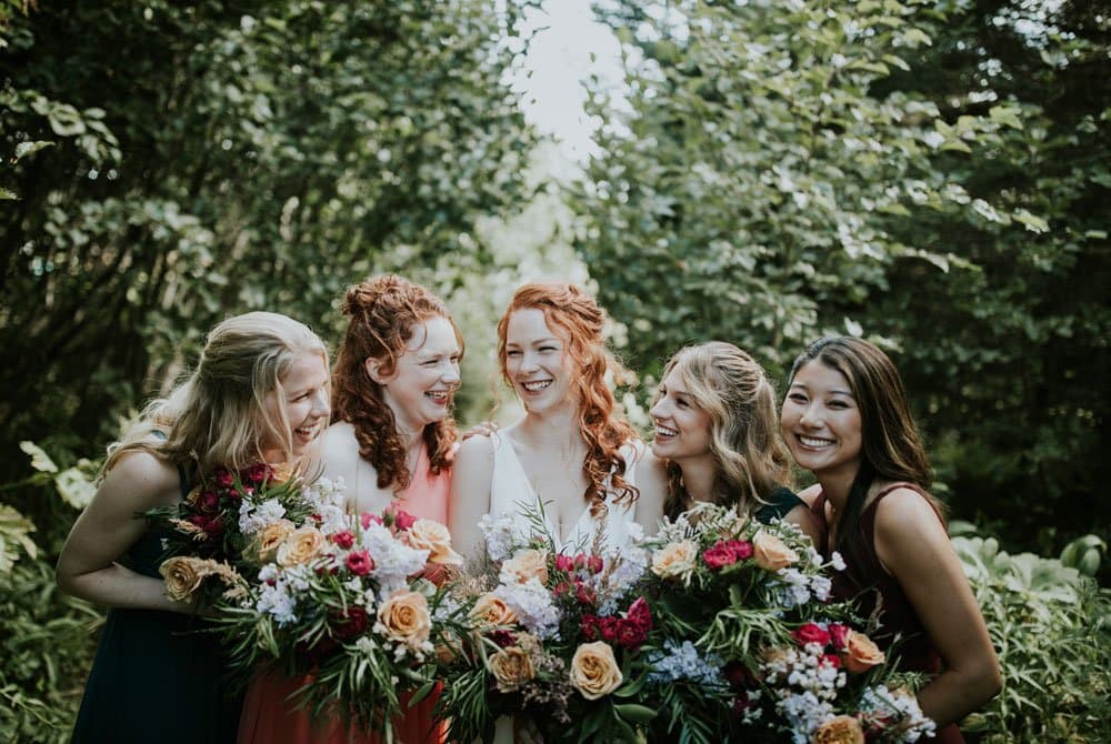 Five bridesmaids with colorful bouquets share a joyful moment in a lush outdoor setting.