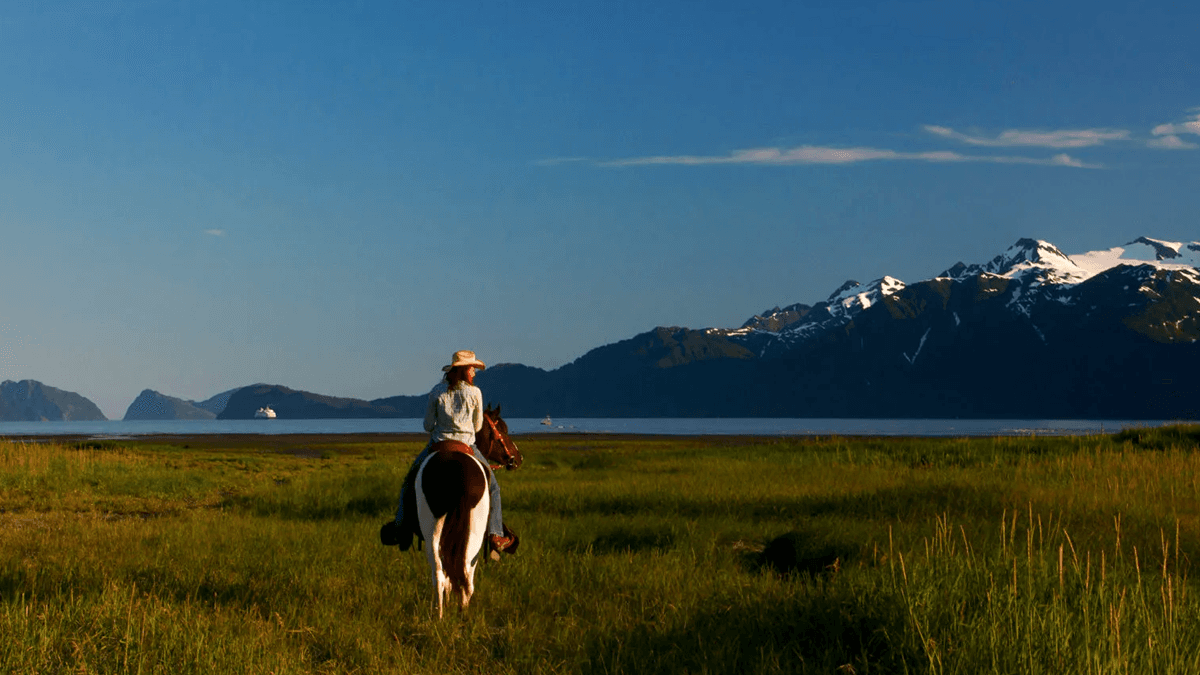 A person on horseback rides through a grassy field with mountains and ocean in the background.