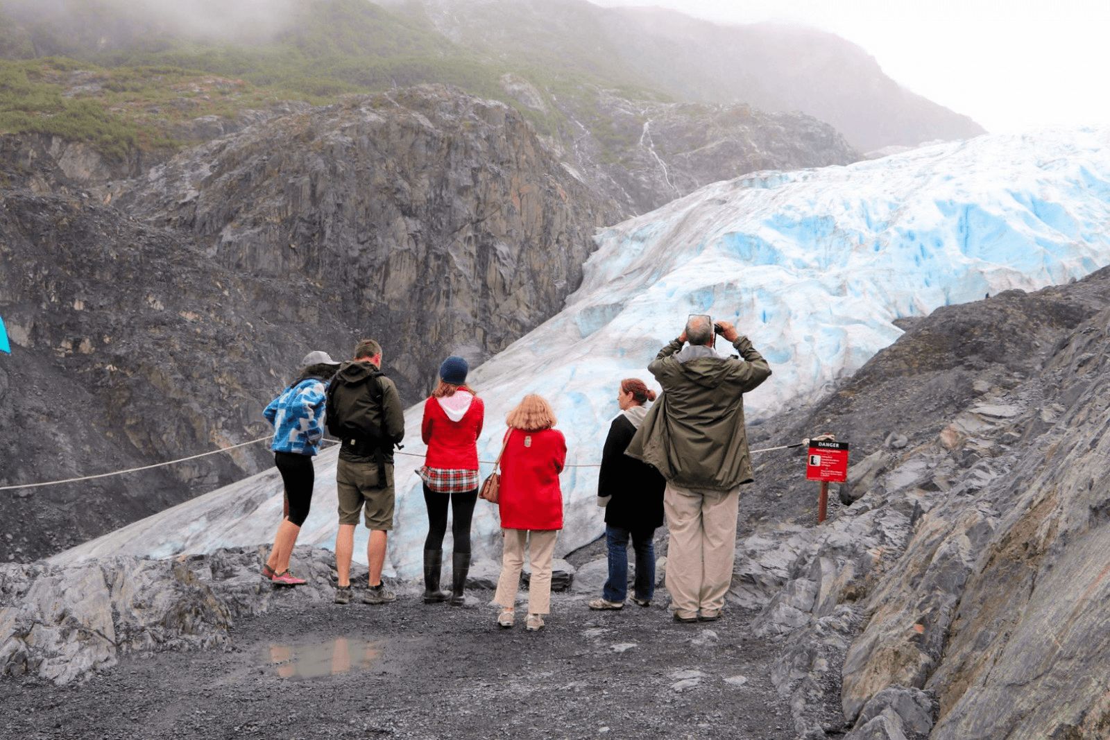 A group of six people stands on rocky terrain, observing a glacier in a misty landscape.